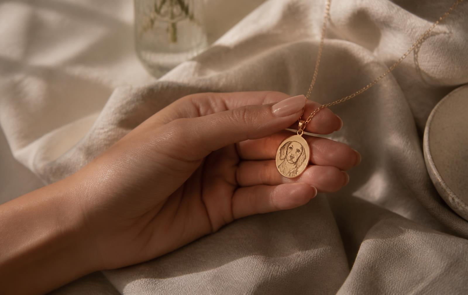 Hand holding a delicate gold pendant engraved with a pet portrait on cream linen